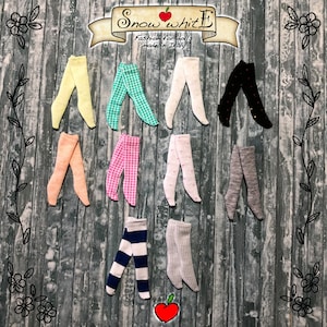 Puede incluir: Una colección de 10 pares de calcetines para muñecas en varios colores y patrones, incluyendo amarillo, rosa, verde, blanco y gris. Los calcetines están dispuestos en dos filas sobre un fondo de madera. El texto "Snow White Fashion for dolls made in Italy" es visible en la parte superior de la imagen.