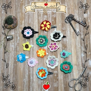 May include: A collection of 12 crocheted flower hair clips in various colors, including pink, blue, green, yellow, and white. The flowers are arranged on a wooden surface with a sign that reads "Snow White Fashion for dolls made in Italy."