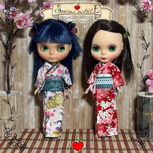 Könnte beinhalten: Zwei Puppen in bunten Kimono-Kleidern. Eine Puppe hat blaue Haare, die andere hat dunkelbraune Haare. Die Puppen haben große grüne Augen und stehen vor einem Holz Hintergrund. Der Text "Snow white" ist oben zu sehen.