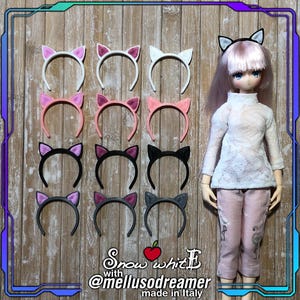 Può includere: Una collezione di cerchietti con orecchie da gatto in vari colori, tra cui rosa, bianco e nero, esposti su una superficie di legno. Una bambola con un top bianco e pantaloni rosa è presente nell'immagine. Il testo "Snow whitE with @mellusodreamer made in Italy" è visibile.