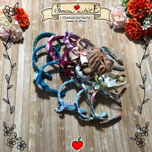 May include: A collection of 10 colorful headbands for dolls, featuring various fabrics and embellishments, including bows, flowers, and glitter. The headbands are arranged on a wooden surface with the text "Snow White Fashion for dolls made in Italy" visible in the background.