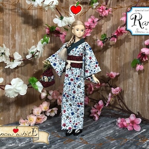 Op de afbeelding: Een pop die een wit en blauw bloemenkimono draagt met een donkerrode band. De pop heeft blond haar en blauwe ogen. De pop houdt een klein mandje vast. De achtergrond is een houten muur met roze en witte bloemen.