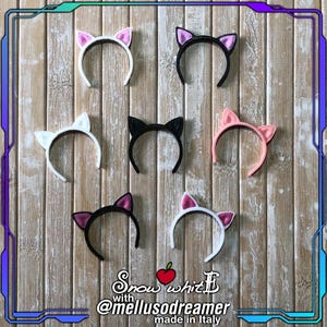 Può includere: Una collezione di cerchietti con orecchie da gatto in vari colori, tra cui bianco, nero e rosa, esposti su uno sfondo di legno. I cerchietti presentano orecchie di diversi colori, alcune con colori interni a contrasto. L'immagine include del testo.