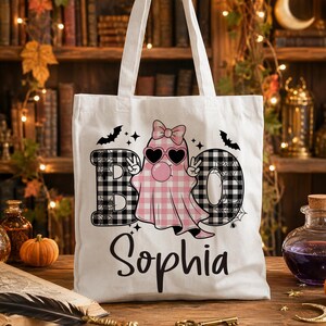 May include: A white canvas tote bag featuring a graphic of a ghost wearing heart-shaped sunglasses and a pink bow, with a pink plaid pattern. The word BOO is printed in black and white plaid, and the name Sophia is written in black script.