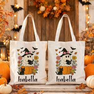 Peut inclure: Deux sacs fourre-tout en toile blanc cassé avec un motif représentant un fantôme portant un chapeau de sorcière et lisant un livre sur une citrouille, accompagné d'un chat noir et de fleurs colorées, avec le nom Isabella imprimé en noir.