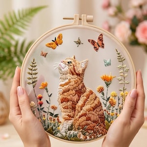 May include: A hand-stitched embroidery hoop featuring a ginger tabby cat gazing upwards. The design includes colorful butterflies, flowers, and foliage. The embroidery is set against a sheer fabric, creating a layered effect within the wooden hoop.