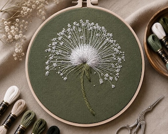 May include: A circular wooden embroidery hoop containing olive green fabric with a hand-stitched white dandelion seed head design. The embroidery features intricate white thread work and small knots, with a green stem extending downward.