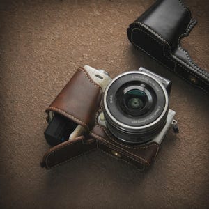 Puede incluir: Una cámara digital plateada y negra con una lente, parcialmente encerrada en una funda de cuero marrón. También se ve una funda de cámara de cuero negro separada. La cámara y las fundas están sobre una superficie marrón texturizada.