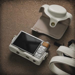 May include: A digital camera encased in a white textured protective cover with a matching detachable top flap. The camera features a silver body, a black screen, and a small tan accent on the top right. A white strap is included in the set.