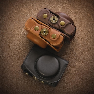May include: Three leather camera cases in shades of brown and black. The cases have brass-coloured hardware and a snap closure. The top two cases are open, revealing the interior. The bottom case is closed with a circular lens opening.