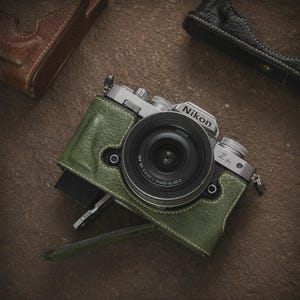 May include: A silver Nikon Z fc camera with a black lens, encased in a green leather case. The camera is positioned on a brown surface, with additional leather cases in brown and black visible in the background.