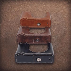 May include: Three camera half cases in light brown, dark brown, and black leather. Each case features a contoured grip, white contrast stitching, a central metal tripod mount screw, and a cutout for the camera lens and battery compartment access.