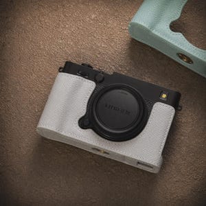 May include: A black camera with a white leather case. The camera has a black lens and a silver button. The case is textured and fits the camera body. A light blue camera case is in the background.