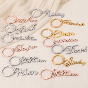 May include: A collection of personalized name keychains in various metallic finishes, including silver, gold, and rose gold. Each keychain features a different name in a cursive font, attached to a circular keyring. The names include "Debora", "Tina", "Elizabeth", and "Jacky".