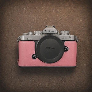 May include: A silver Nikon Z fc camera with a black lens cap and a pink leather case. The camera has the Nikon logo on the top and lens cap. The pink case wraps around the camera body.