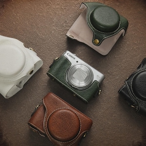 Op de afbeelding: Een verzameling leren camerahoesjes in verschillende kleuren, waaronder wit, groen, bruin en zwart. Een zilveren Canon-camera is zichtbaar, gedeeltelijk omhuld door een groene leren hoes. De hoesjes hebben een cirkelvormig ontwerp.