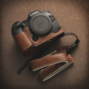 K&ouml;nnte beinhalten: Eine schwarze Canon EOS R6 Kamera mit brauner Lederh&uuml;lle und Objektivdeckel. Die Kamera hat einen passenden braunen Lederriemen. H&uuml;lle und Riemen sind mit wei&szlig;em Faden gen&auml;ht. Die Kamera liegt auf einer braunen Oberfl&auml;che.