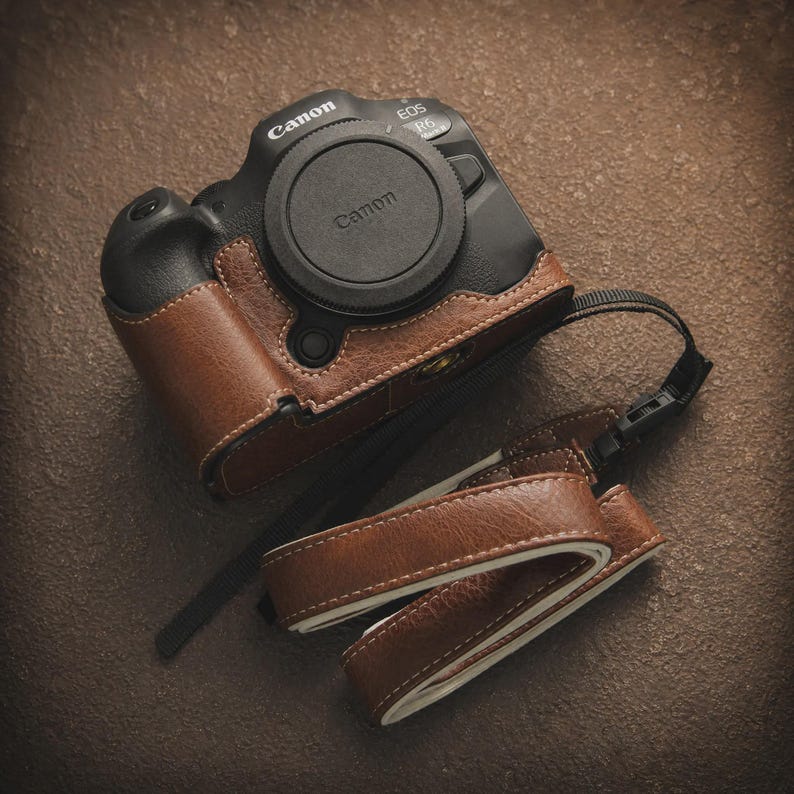 K&ouml;nnte beinhalten: Eine schwarze Canon EOS R6 Kamera mit brauner Lederh&uuml;lle und Objektivdeckel. Die Kamera hat einen passenden braunen Lederriemen. H&uuml;lle und Riemen sind mit wei&szlig;em Faden gen&auml;ht. Die Kamera liegt auf einer braunen Oberfl&auml;che.