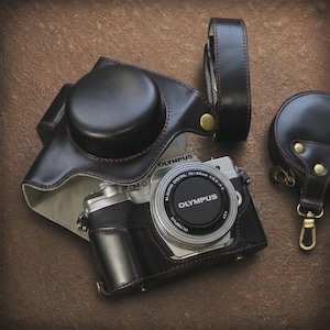 May include: A silver Olympus OM-D camera with a black lens cap and M.Zuiko Digital 14-42mm lens, encased in a black leather half-case with red stitching. Includes a matching black leather camera strap and a small circular black leather lens cap pouch.