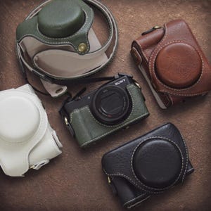 May include: A black Sony camera with a green leather-style case, surrounded by three additional camera cases in white, olive green, and brown. Each case features a circular lens cover, visible stitching, and metallic snap buttons for secure closure.