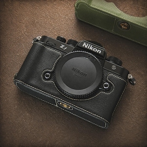 Może przedstawiać: Czarny aparat Nikon Z f z osłoną obiektywu, w czarnym skórzanym etui z białymi przeszyciami. Zielone etui na aparat znajduje się w tle. Aparat posiada logo Nikon i napis "Z f".