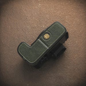 K&ouml;nnte beinhalten: Eine gr&uuml;ne Kameratasche aus Leder mit wei&szlig;en N&auml;hten. Die Tasche bedeckt einen schwarzen Kamerak&ouml;rper. Die Tasche hat einen goldfarbenen Knopf und eine strukturierte Oberfl&auml;che. Die Kameratasche dient zum Schutz des Kamerageh&auml;uses.