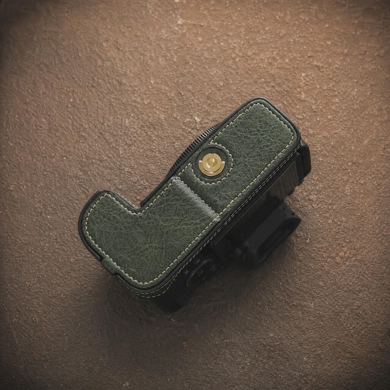 K&ouml;nnte beinhalten: Eine gr&uuml;ne Kameratasche aus Leder mit wei&szlig;en N&auml;hten. Die Tasche bedeckt einen schwarzen Kamerak&ouml;rper. Die Tasche hat einen goldfarbenen Knopf und eine strukturierte Oberfl&auml;che. Die Kameratasche dient zum Schutz des Kamerageh&auml;uses.