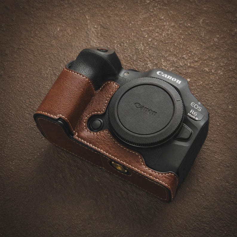 K&ouml;nnte beinhalten: Eine braune Leder-Kameratasche mit wei&szlig;en N&auml;hten, passend f&uuml;r eine Canon EOS R6 Mark II Kamera. Der schwarze Objektivdeckel tr&auml;gt das Canon-Logo. Die Tasche umschlie&szlig;t das Kamerageh&auml;use und bietet Schutz und ein stilvolles Aussehen.