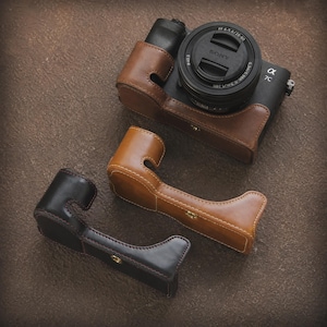 Peut inclure: Un appareil photo Sony Alpha 7C noir avec un étui en cuir marron et deux poignées en cuir supplémentaires. L'objectif de l'appareil photo est noir avec du texte blanc. Les étuis et poignées en cuir sont cousus avec du fil blanc.