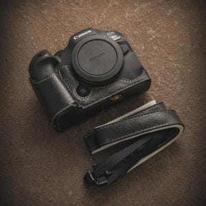 K&ouml;nnte beinhalten: Schwarze Kameratasche und -riemen aus Leder. Die Tasche ist f&uuml;r eine Canon EOS R6 Kamera konzipiert und hat wei&szlig;e N&auml;hte. Der Riemen ist aus schwarzem Leder mit einem schwarz-wei&szlig; gewebten Nylonabschnitt. Das Canon-Logo ist sichtbar.