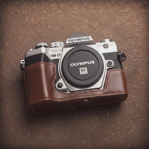 Peut inclure: Un appareil photo numérique Olympus OM-D doté d'un boîtier argenté et noir avec un étui de protection en cuir marron. L'appareil possède un bouchon d'objectif noir avec le logo Olympus et le symbole Micro Four Thirds imprimés en blanc.