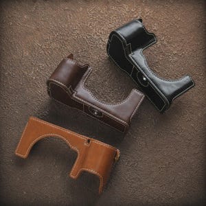 May include: Three leather camera cases in brown, black, and tan. Each case is stitched with white thread and features a form-fitting design. The cases are designed to protect the camera body.