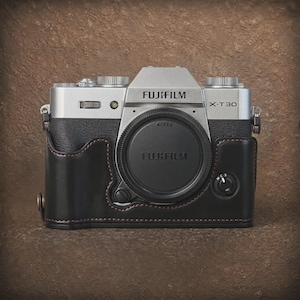 May include: A silver and black Fujifilm X-T30 mirrorless camera body featuring a black lens cap with the Fujifilm logo. The camera is encased in a black leather half-case with visible red stitching, metal strap lugs, and a focus mode selector dial.