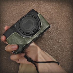 Puede incluir: Una cámara digital negra con una funda de cuero verde. La lente de la cámara está centrada y la funda tiene un diseño cosido. Se adjunta una correa de muñeca negra. La cámara se sostiene en una mano.