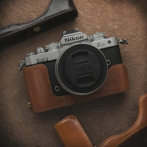 May include: A Nikon Z fc camera with a black lens cap that reads "NIKKOR". The camera is in a brown leather case. Additional brown leather camera cases are in the background.