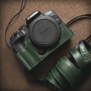 Puede incluir: Un cuerpo de cámara sin espejo Canon EOS R10 negro con tapa de objetivo, dentro de una funda de media carcasa de cuero verde oscuro. Una correa de cámara de cuero verde oscuro a juego está enrollada al lado, con costuras visibles.
