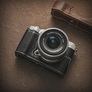 Könnte beinhalten: Eine silberne und schwarze Digitalkamera mit einem Objektiv mit der Aufschrift "FUJINON ASPHERICAL LENS". Die Kamera befindet sich in einer schwarzen Ledertasche mit weißen Nähten. Im Hintergrund befindet sich eine braune Ledertasche.