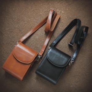 May include: Two leather crossbody bags. One is a light brown color, the other is black. Both have a rectangular shape with a curved flap closure and adjustable straps. The bags are displayed on a textured brown surface.