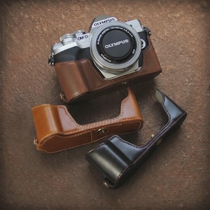 Może przedstawiać: Aparat cyfrowy Olympus OM-D ze srebrno-czarnym korpusem i dekielkiem obiektywu, wyposażony w brązowy skórzany pokrowiec typu half-case. Poniżej aparatu znajdują się dwa dodatkowe skórzane pokrowce w kolorze jasnobrązowym i ciemnobrązowym.