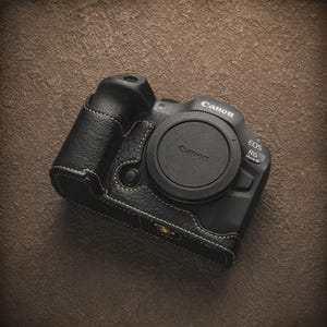 K&ouml;nnte beinhalten: Eine schwarze Canon EOS R6 Mark II Kamera mit Objektivdeckel, in einem schwarzen Lederetui mit wei&szlig;en N&auml;hten. Die Kamera liegt auf einer strukturierten braunen Oberfl&auml;che. Das Canon-Logo ist sichtbar.