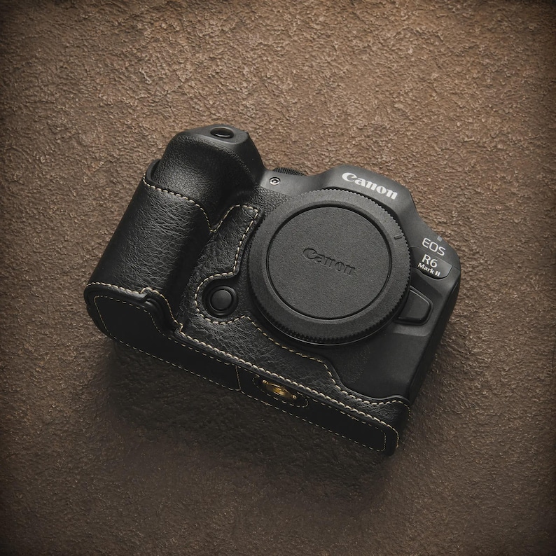 K&ouml;nnte beinhalten: Eine schwarze Canon EOS R6 Mark II Kamera mit Objektivdeckel, in einem schwarzen Lederetui mit wei&szlig;en N&auml;hten. Die Kamera liegt auf einer strukturierten braunen Oberfl&auml;che. Das Canon-Logo ist sichtbar.
