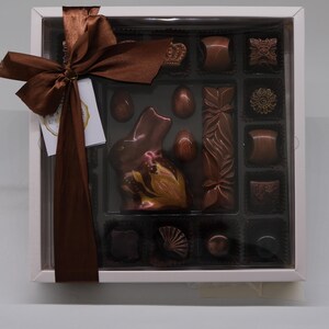 Può includere: Una scatola di cioccolatini assortiti, tra cui un coniglietto di cioccolato, uova e varie forme. I cioccolatini sono in tonalità marroni e sono disposti in una scatola bianca con coperchio trasparente. Un nastro marrone adorna la scatola.