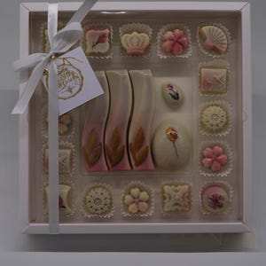 Peut inclure: Une boîte blanche de chocolats assortis avec un couvercle transparent, attachée avec un ruban blanc. Les chocolats sont décorés de motifs floraux et abstraits roses, blancs et bruns. Une petite étiquette avec du texte doré est attachée au ruban.