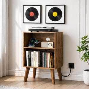 May include: A mid-century modern style wooden record player stand with a two-shelf design. The stand features a turntable, framed vinyl records, and a small radio. A smartphone and smartwatch are on the top shelf. The stand is brown with angled legs.