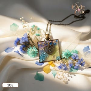 May include: A square pendant necklace with a bronze chain. The pendant features a heart-shaped charm and a colorful design with blue, purple, and yellow elements. The necklace is displayed on a white satin surface with blue and green flowers.