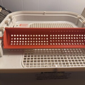 May include: A bright orange, rectangular, perforated plastic tray with rounded ends. The tray is positioned inside a white plastic container with a grid-like pattern. The container has a white lid and a white base.