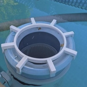 May include: A light blue pool skimmer with a white, star-shaped handle. The skimmer is partially submerged in clear blue water, with a gray hose visible in the background. The skimmer is designed to remove debris from the pool.