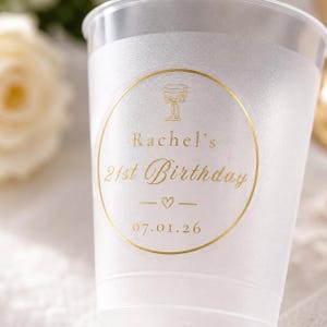 Op de afbeelding: Een doorzichtige plastic beker met een gouden folie label. Het label leest "Rachel's 21st Birthday" met een champagneglas icoon, een hartje en de datum 07.01.26. De beker heeft een matte uitstraling.