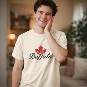 Puede incluir: Camiseta color crema con un diseño de hoja de arce roja sobre la palabra "Buffalo" en fuente cursiva. La camiseta es de manga corta y cuello redondo.
