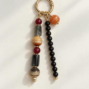 May include: A gold-toned metal ring keychain featuring two dangling strands of beads. One strand has alternating red, gray, tan, and black cylindrical and spherical beads. The second strand consists of small black beads and one large orange spherical bead.